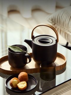 Une théière noire et une tasse sur un plateau en bois avec une assiette de muffins sur une table en verre à côté d'un canapé dans une pièce ensoleillée.