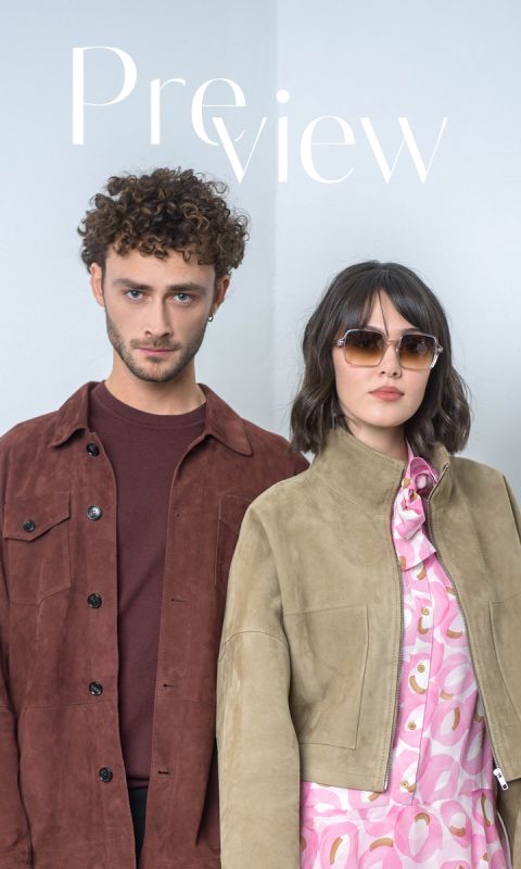 Un homme portant une veste marron et une femme portant des lunettes de soleil et une veste marron clair sur une robe à motifs roses se tiennent côte à côte devant un fond clair et uni.