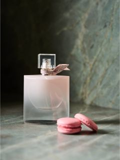 Un flacon de parfum rose avec un nœud est posé à côté de deux macarons roses sur une surface en marbre.