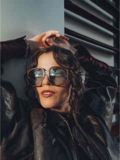 Une femme avec de grandes lunettes de soleil et une veste en cuir noire se penche en arrière avec les mains derrière la tête, la lumière du soleil se reflétant dans ses lunettes.