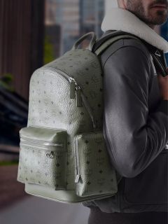 Une personne vêtue d'une veste noire porte un grand sac à dos MCM vert olive texturé avec des poches zippées et se tient à l'extérieur dans un environnement urbain.