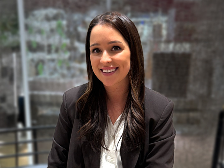 Une femme aux longs cheveux noirs, portant un blazer noir et une chemise blanche, sourit assise dans une maison, avec un arrière-plan flou.