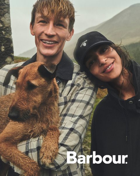 Un homme souriant avec un chien marron se tient à côté d'une femme souriante avec un bonnet, tous deux à l'extérieur avec des collines en arrière-plan. Le logo Barbour est visible.