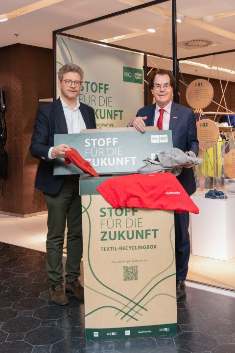 Deux hommes se tiennent à côté d'un conteneur de recyclage textile portant l'inscription "Des tissus pour l'avenir" et tiennent des vêtements dans un intérieur moderne avec des bannières d'information.