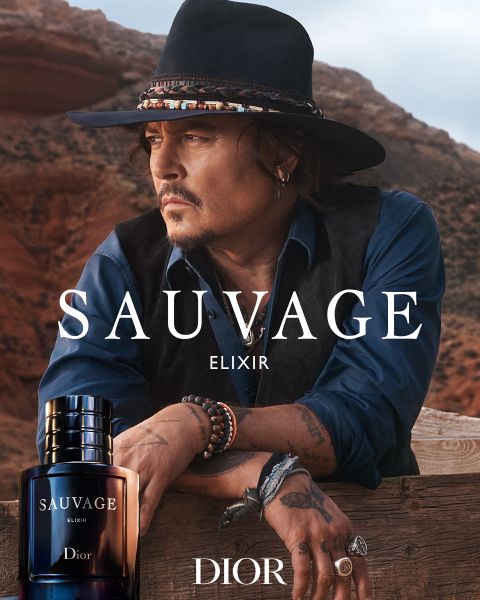 Un homme avec un chapeau et un gilet s'appuie sur une balustrade en bois dans le désert, au premier plan un flacon du parfum Sauvage Elixir Dior avec logo.