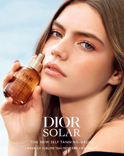 Une femme au teint éclatant tient un flacon de gouttes autobronzantes Dior Solar près de son visage, sur lequel on peut lire en blanc le nom du produit et le slogan.