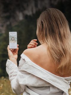 Une femme aux cheveux longs et à la chemise blanche épaules nues tient un produit de soin de la peau à l'extérieur et regarde vers un arrière-plan forestier flou.