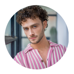 Homme aux cheveux bruns bouclés et aux yeux bleus, portant une chemise rayée rose et blanche, regardant la caméra à l'intérieur près d'une fenêtre.