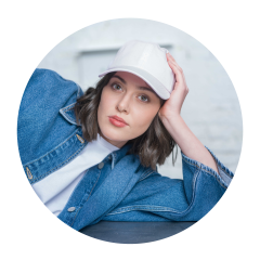 Femme avec un bonnet blanc et une veste en jean, s'appuyant sur son bras et regardant l'appareil photo avec un regard neutre.