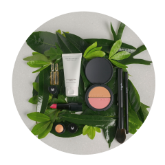 Produits de maquillage, notamment du blush, du rouge à lèvres, des pinceaux et des crèmes, disposés en cercle sur des feuilles vertes sur un fond clair.