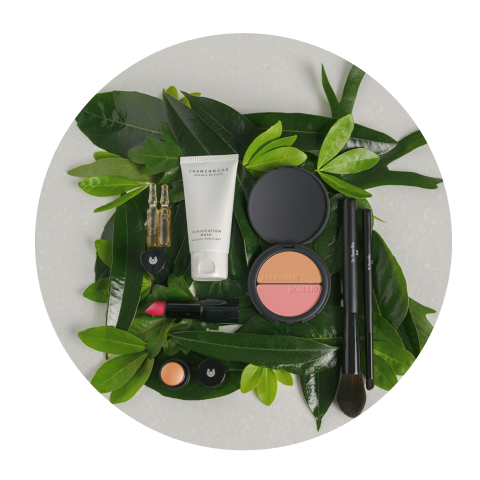 Produits de maquillage, notamment du blush, du rouge à lèvres, des pinceaux et des crèmes, disposés en cercle sur des feuilles vertes sur un fond clair.