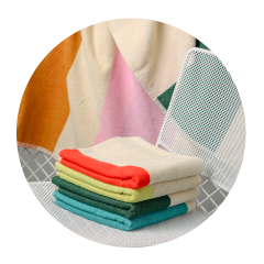 Une pile de serviettes colorées et pliées sur une chaise en filet blanche, avec une couverture à motifs drapée en arrière-plan.