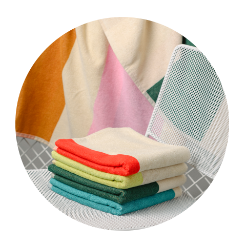 Une pile de serviettes colorées et pliées sur une chaise en filet blanche, avec une couverture à motifs drapée en arrière-plan.