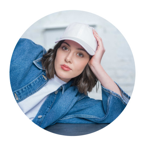 Femme avec un bonnet blanc et une veste en jean, s'appuyant sur son bras et regardant l'appareil photo avec un regard neutre.