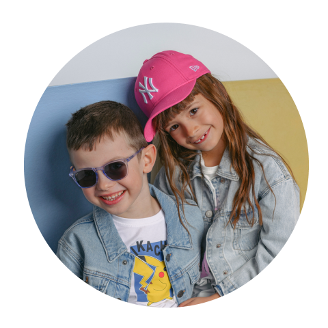 Deux enfants souriants en vestes en jean ; le garçon porte des lunettes de soleil, la fille une casquette de baseball rose et appuie sa tête contre la sienne.