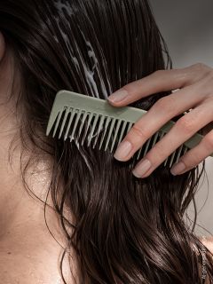 Une personne peigne des cheveux foncés et mouillés avec un peigne à dents larges, répartissant l'après-shampoing dans les mèches.