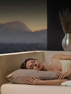 Une femme dort sur le côté dans un lit avec un oreiller beige, à côté d'une fenêtre qui montre un paysage de montagne flou au crépuscule.