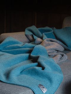 Une couverture en laine bleue avec un dessous gris est drapée sur un canapé gris, avec une étiquette en tissu visible au premier plan.