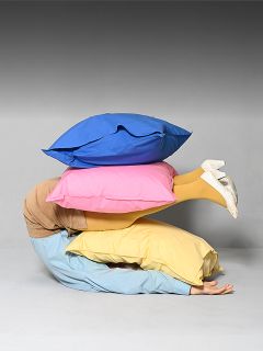 Une personne est allongée, recroquevillée sur le sol, recouverte de trois grands coussins colorés, desquels ne dépassent que ses jambes vêtues de jaune et ses chaussures blanches.