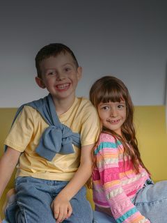 Deux enfants souriants, un garçon et une fille, sont assis ensemble sur un canapé jaune. Le garçon porte une chemise jaune, la fille un pull à rayures multicolores.