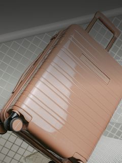 Une valise rigide brillante, brun clair, avec des roulettes et une poignée extensible est posée sur une surface carrelée.