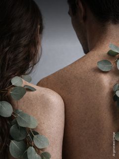 Deux personnes aux dos nus et couverts de taches de rousseur se tiennent près l'une de l'autre, des feuilles d'eucalyptus drapées sur les épaules, devant un fond neutre.
