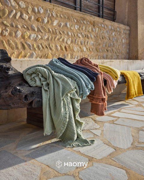 Des jetés de lit colorés et texturés drapés sur un banc en bois rustique à l'extérieur, avec un mur de pierre et un trottoir en arrière-plan. Logo Naomy en bas.