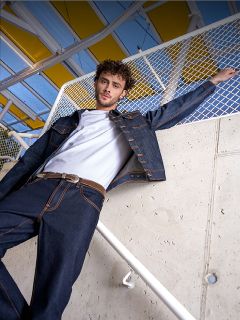 Un homme en t-shirt blanc, veste en jean foncée et jean se tient à une balustrade appuyée contre un mur en béton et une clôture, surmontée d'un auvent jaune.