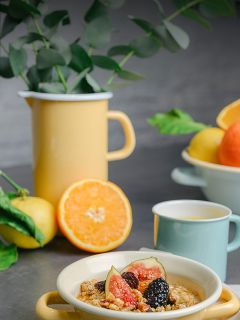 Un bol de céréales, de figues et de baies, un pichet jaune avec des branches vertes, des agrumes et une tasse bleu clair sur une table grise.