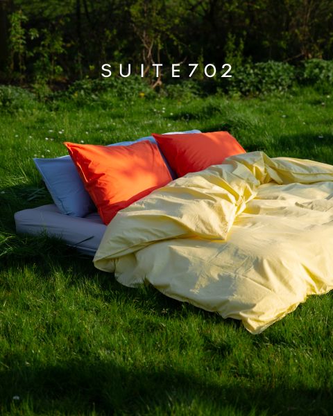 Un lit avec des coussins orange vif et rouges et une couette jaune se trouve à l'extérieur sur de l'herbe verte au soleil. Au-dessus, il est écrit « SUITE702 ».