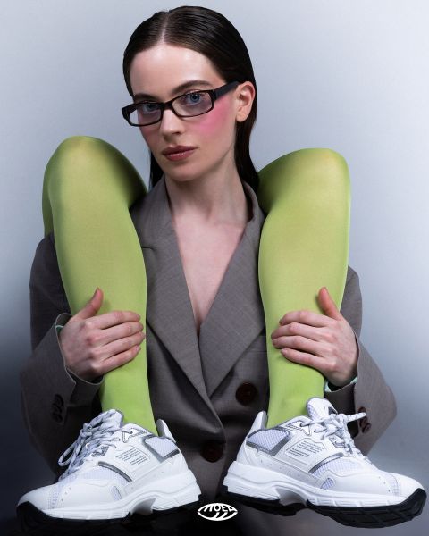 Une femme portant un blazer gris, des lunettes et des baskets blanches, tient ses jambes sur ses épaules avec des chaussettes vert vif, devant un fond uni.
