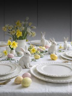 Table de Pâques dressée avec des assiettes blanches, des figurines de lapins en céramique, des œufs de couleur pastel et un vase de jonquilles jaunes sur une nappe blanche.