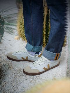 Une personne en jeans retroussés se tient sur un sol sablonneux près de cactus, portant des baskets blanches avec des accents beiges et marrons et un logo "V" sur le côté.