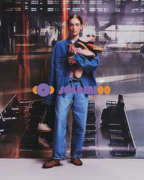 Une femme en veste en jean et en jean se tient dans un terminal d'aéroport et tient plusieurs paires de baskets dans ses bras.