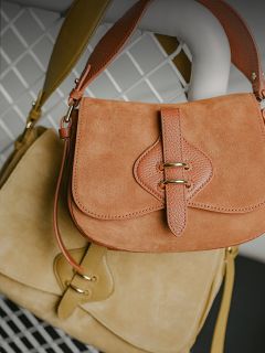 Deux sacs à main en daim, un marron clair et un beige, avec des lanières en cuir et des fermoirs dorés, suspendus sur un fond de treillis métallique blanc.