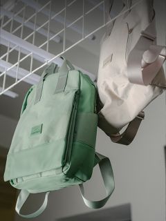 Deux sacs à dos, un vert menthe et un beige, sont suspendus à une structure métallique blanche dans une pièce bien éclairée.