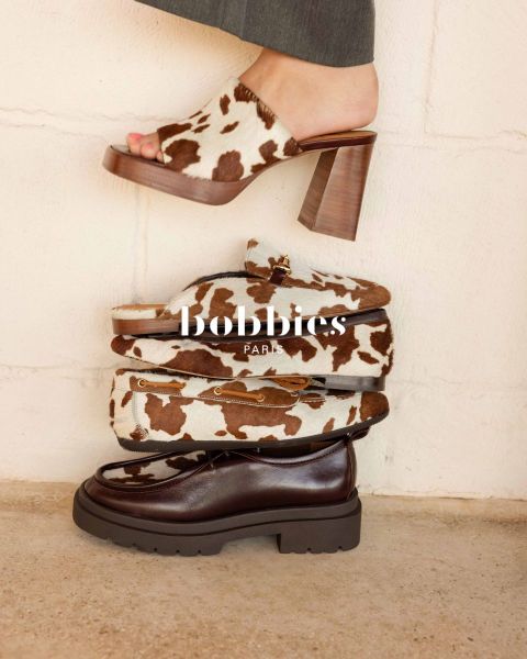 Quatre chaussures à la mode, dont des talons à imprimé vache, des mocassins et une chaussure épaisse marron foncé, empilées devant un mur clair avec une superposition de texte "Bobbies Paris".