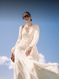Une femme en robe blanche flottante et grandes lunettes de soleil sourit à l'extérieur devant un ciel bleu avec quelques nuages et porte un sac marron.