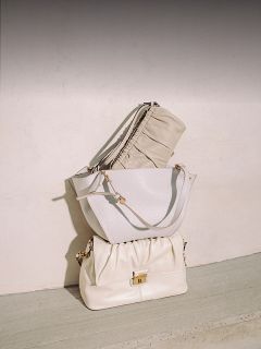 Trois élégants sacs à main beiges et blancs sont empilés devant un mur clair et un sol en béton, et disposés avec soin à la lumière naturelle du soleil.