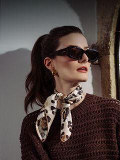 Femme portant des lunettes de soleil foncées, des boucles d'oreilles dorées, un foulard à motif léopard et un pull texturé marron, posant près d'une fenêtre avec de la lumière sur son visage.