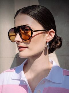 Une femme aux cheveux bruns, coiffés en un chignon bas, porte de grandes lunettes de soleil couleur ambre, des boucles d'oreilles argentées et une chemise rose et blanche à col, baignée de soleil.