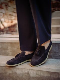 Personne en chaussures en daim foncé à glands et pantalon foncé, debout sur un escalier en béton à l'extérieur avec un arrière-plan flou de mur de briques.