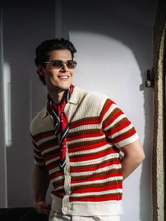 Un homme souriant avec des lunettes de soleil, une chemise rayée rouge et blanche et une écharpe se tient au soleil devant un mur blanc.