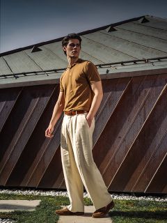 Un homme en polo marron et pantalon ample couleur crème sort près d'un bâtiment moderne avec un toit métallique incliné.