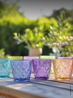 Quatre gobelets en verre structurés et colorés sont posés sur une table en bois à l'extérieur, avec des plantes vertes floues et la lumière du soleil en arrière-plan.