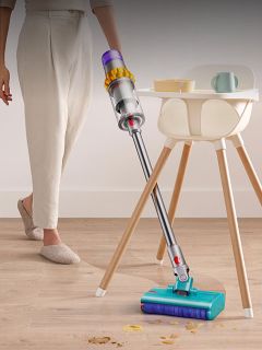 Une personne utilise un aspirateur sans fil pour débarrasser un plancher en bois des miettes près d'une chaise haute et d'une petite table avec de la vaisselle.