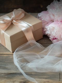 Boîte cadeau avec un ruban rose tendre à côté de pivoines rose tendre et de tissu blanc sur une surface en bois.