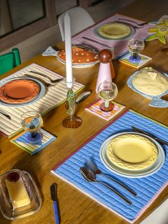 Une table à manger en bois avec des assiettes colorées, des sets de table matelassés, des verres, des couverts et des bougies dans un arrangement lumineux et éclectique.