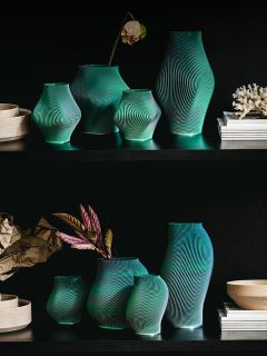 Sur des étagères noires se trouve une collection de vases verts avec des lignes blanches ondulées, certains avec des fleurs, entourés de livres et d'objets de décoration.