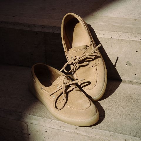 Une paire de chaussures bateau beiges en daim avec des lacets, posées sur des marches en béton sous un soleil partiel.